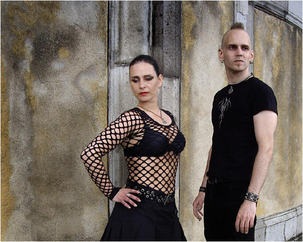 Mantus-bändin kaksi jäsentä seisoo betoniseinän edustalla mustiin vaatteisiin pukeutuneina. Vasemmanpuoleinen on mustahiuksinen goottinainen, jolla musta hame ja verkkopaita yllään. Naisen oikealla puolen mustaan hihattomaan paitaan ja mustiin housu