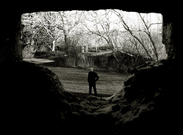 Valokuva mustiin pukeutuneesta lyhythiuksisesat miehestä. Hän seisoo nurmikolla kuolleiden puiden edustalla. Kuva otettu muurin yläpuolelta, sillä etualalla näkyy suuria kivilohkareita.