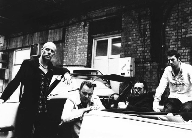 Harmaasävyinen ja ylivalottunut valokuva Ausgang-bändin kokoonpanosta, johon kuuluu neljä mieshenkilöä. Miehet ovat avoauton ympärillä ja sen istuimilla kivitiilitalojen edustalla.