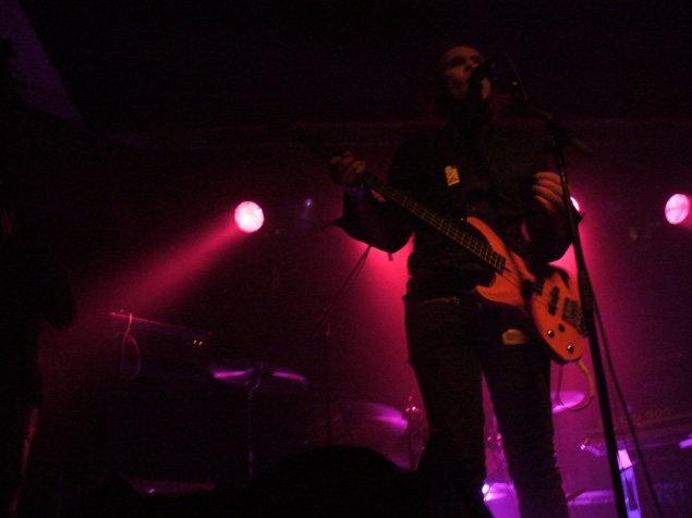 Valokuva Waltari-bändin laulaja Hatakasta, joka soittaa bassoa samalla kun laulaa mikrofoniin. Miehellä mustat vaatteet. Taustalla spottivaloja, joista punaista ja violettia valoa lankeaa lavalle.