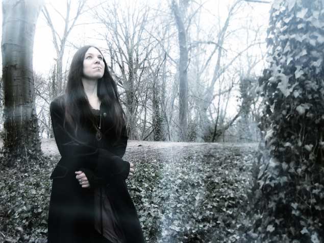 Mustaan pitkään takkiin pukeutunut tummahiuksinen nainen seisoo elottomien ja lehdettömien puiden seassa keskellä korpea. Nainen katsoo ylöspäin. Hänellä on alahuulessa huulikoru.