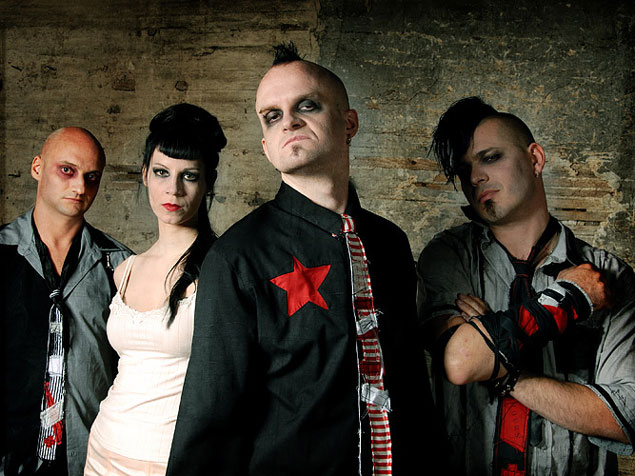 Metallspürhunden kokoonpanossa kolme mistä ja yksi valkoiseen mekkoon pukeutunut mustahiuksinen nainen. Henkilöt seisovat rivissä rapautunutta puuseinää vasten. Miehillä kalpeat ihot ja omalaatuiset vaatteet yllään.