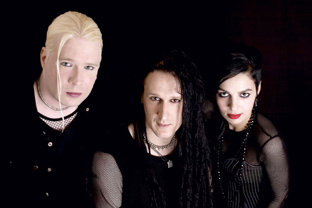 Kolme ihmistä, kaksi miestä ja nainen, seisovat pikimustaa taustaa vasten pukeutuneina mustiin vaatteisiin. Heistä kullakin pitkät hiukset. Vasemman laidan miehellä valkoinen tukka, muilla mustat hiukset. Naisella punatut huulet.