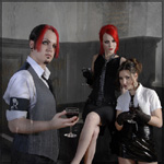 Kolme ihmistä harmaata taustaa vasten. Vasemmassa laidassa mies, oikealla kaksi istuvaa naista. Keskimmäisellä naisella tulenpunaiset hiukset, kuten miehelläkin. Miehellä on tummanharmaa hihanauha käsivarressaan.