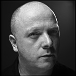 Harmaasävyinen valokuva kaljupäisestä Ronan Harriksesta, VNV Nationin miehestä. Kuva on tummanharmaata taustaa vasten. Miehen kasvot ovat ilmeettömät.