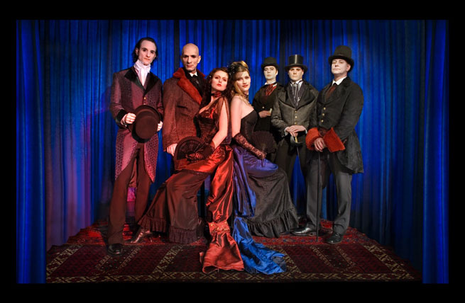 Aeternitaksen jäsenet koristeellisten mattojen päällä sähkönsinistä seinäkangasta vasten. Keskimmäisenä on kaksi naista, jotka istuvat selätysten. Naisten ympärillä viisi miestä, joista kaikilla vanhanaikaiset ja mustat pvuut yllään.