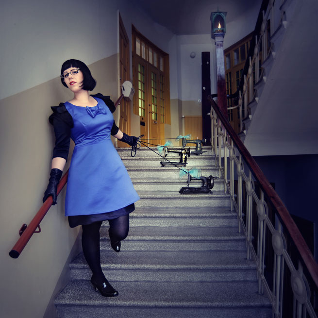 Valokuva siniseen mekkoon pukeutuneesta mustahiuksisesta naisesta, joka raahaa talutushihnassaan vanhanaikaisia ompelukoneita porraskäytävässä.