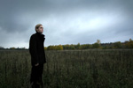 Valokuva pellolla seisovasta pitkähiuksisesta miehestä, jolla mustat vaatteet yllään. Hän seisoo vasemmalla katsellen oikealle. Kuvan yläosassa sininen taivas, joka pilvinen.