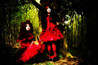 Kaksi punaisiin goottilolitamekkoihin pukeutunutta naista synkässä ja tummanvihreässä luonnossa nojailemassa puuta vasten. Kummallakin naisella runsaasti vaatetusta yllään.