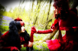 Punaisiin goottiloliltapukuihin sonnustautuneita naisia kaksi kappaletta. He pitävät toisiaan kädestä kiinni. Taustalla näkyy vihreää ja ylivalottunutta, voimakaskontrastista viherikköä.