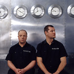 Valokuva VNV Nationin kahdesta mustiin pukeutuneesta jäsenestä, miehestä, jotka istuvat teräksistä seinää vasten. Seinällä on runsaasti kelloja, jotka näyttävät kaikki eri aikaa. Kuvan jäsenillä lyhythihaiset mustat paidat ja lyhyet hiukset.