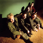 Front Line Assembly -bändin promokuva, jossa viisi miestä istuu lattialla. Taustalla varjoja ja vihertävää seinää.