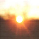 Valokuva auringonlaskusta tai -noususta. Kuvassa näkyy häikäisevä kellertävä valopallo ja epätarkkaa maaperää tai vettä alaosassa. Yläosassa kellertävää puhkipalanutta aluetta.