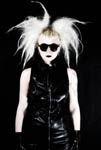 Valokuva vaaleahiuksisesta goottinaisesta, jolla pörröinen tukka ja musta nahka-asu. Naisella mustat aurinkolasit silmillä. Kuvan tausta on musta.