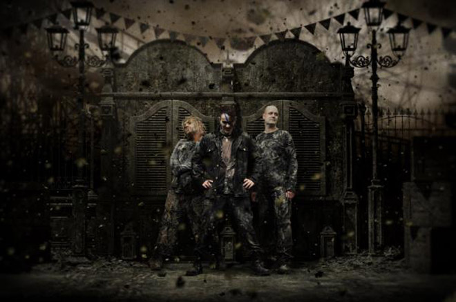 Ryhmäkuva Project Pitchforkin jäsenistä, jotka seisovat rivissä mustiin pukeutuneena elotonta ja nuhjuista taustaa vasten. Ilma on täynnä pölyä ja tuhkaa.