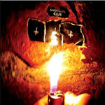 Valokuva kynttilästä, joka palaa kiven edessä ja tuottaa voimakkaan keltaista väriä. Kiven päällä mustia laattoja, joissa risti ylösalaisin.