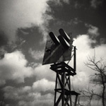 Harmaasävyinen valokuva jonkinlaisesta puurakennelmasta, tornisa, jossa putkia. Kuvan taustalla näkyy pilvinen taivas ja kuolleen puun oksistoa.