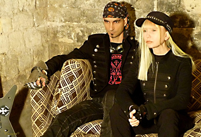 Kaksi mustiin pukeutunutta muusikkoa istuu sohvalla. Vasemmalla on mies, jolla bandanna päässä. Oikealla lippispäinen, vaaleahiuksinen nainen.