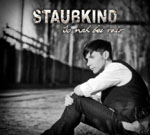 Harmaasävyinen valokuva miehestä istumassa maassa aidan vierustalla. Kuvan yläosassa suurella fonttikoolla Staubkind-logo.