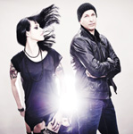 Kuva miehestä ja naisesta, jotka seisovat vaaleanharmaata taustaa vasten. Molemmilla mustat vaatteet. Naisen hiukset liehuvat.