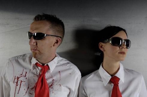 Suicide Commando -yhtyeen jäsenet, eli yksi mies ja yksi nainen, seisovat vaaleata seinää vasten. Molemmilla yllään identtiset vaatteet ja asusteet: punainen solmio, kauluspaita, mustat aurinkolasit. Vasemmassa laidassa näkyvällä miehellä kuitenk