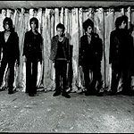 Mustavalkoinen ryhmäkuva Dir En Grey -bändin jäsenistä, jotka seisovat jonkinlaista vaaleata seinää vasten. Miehillä mustat vaatteet.