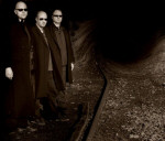 Tummasävyinen valokuva 32Crash-yhtyeen jäsenistä, jotka seisovat mustat vaatteet yllään ja mustat aurinkolasit silmillä kaivostunnelin seinän vierustalla. Tunnelin lattialla kulkee raiteet.