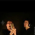 Amnistia-bändin jäsenet promokuvassa. Kuvassa kaksi lyhythiuksista miestä, joista kummallakin silmälasit. Vasemmanpuoleinen huutaa mikrofoniin ja oikeanpuoleisella korvakuulokkeet päässä.