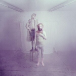Pastellinsävyinen, violetilla ja valkoisella päällystetty valokuva kahdesta ihmishahmosta seisomassa matalassa huoneessa.