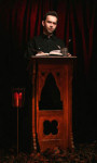 Lyhythiuksinen ja vaaleaihoinen mieshenkilö seisoo tummasävyisessä valokuvassa jonkinlaisen puhujakorokkeen takana. Miehellä on korokkeella avattuna kirja ja kädessään jonkinlainen kynä.