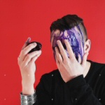 Valokuva miehestä, jolla musta irokeesi päässä ja violettia mustetta kasvojen edessä. Tausta on verenpunainen.