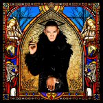 Valokuva koristeellisesta, värikkäästä mosaiikkilasiseinästä, jota vasten kuvassa mustaan karvapukuun sonnustautunut mies.