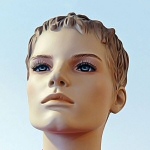 Valokuva nuken päästä kuvattuna vaaleansinistä taustaa vasten. Nuken kasvot ovat miehekkäät, tukka vaaleanruskea.