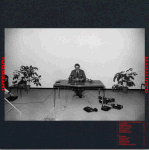 Tummansinistä taustaa vasten asetettu harmaasävyinen valokuva pöydän ääressä istuvasta ihmisestä.