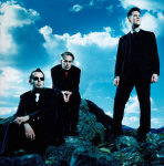 Kolme Covenant-yhtyeen jäsentä poseeraa sinisävyisessä valokuvassa mustiin vaatteisiin pukeutuneina. Taustalla pilvinen ja sininen taivas, miehet seisovat / istuvat kivikossa.