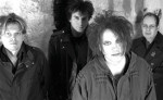 Mustavalkoinen valokuva The Cure -yhtyeen neljästä miesjäsenestä. He seisovat vaaleata seinää vasten. Etummainen mies näyttää naiselta.