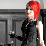 Kirkkaanpunaiset hiukset omaava mustiin pukeutunut naishenkilö seisoo viinilasi kädessään musta&ndash;valkean koristeellisen seinän edustalla.