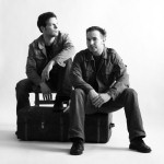 Harmaasävyinen valokuva Endanger-bändin kahdesta miehestä, jotka istuvat mustan matkalaukun päällä valkoista ja vaaleanharmaata taustaa vasten. Molemmilla miehistä lyhyet hiukset ja mustat housut sekä kengät.