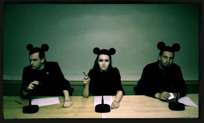 Forgotten Sunrise, joka on virolainen goottiryhmä, hämyisässä valokuvassa. Keskimmäisenä nainen, jonka molemmilla puolilla yksi mies. Jokaisella heistä päässä mustat mikkihiirimäiset korvat. He istuvat pöydän edessä, valkoiset paperit edessään.