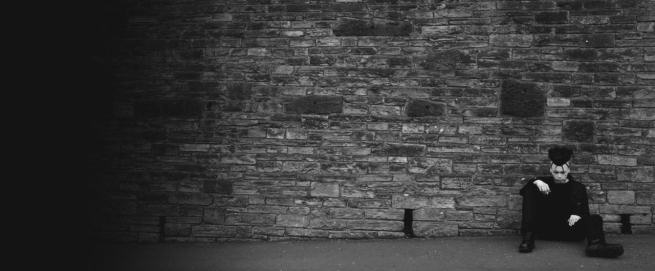 Audioclav.1.1:n Tony Young istuu harmaasävyisessä bändikuvassa kiviseinää vasten asvalttitiellä. Kuvan vasen laita hämärän peitossa. Miehellä musta irokeesimainen letti päässä ja mustat vaatteet, vitivalkoinen iho.