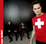 elegant MACHINERY -bändin kolme miestä promokuvassa. Heistä kaksi mustiin pukeutunutta istuu taustalla mustaa kiiltäväpintaista seinää vasten punaisilla tuoleilla. Oikeassa laidassa seisoo punapaitainen mies, jonka paidan keskellä suuri valkoinen