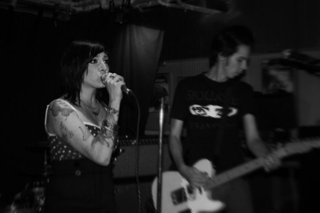 Harmaasävyinen valokuva Angelica Kultin vokalistista, joka on mustahiuksinen nainen ja joka laulaa mikrofoniin katsellen suoraan eteensä. Naisen takana näkyy epätarkasti varjojen kätköissä valkoista kitaraa/bassoa soittava miesmuusikko, jolla musta