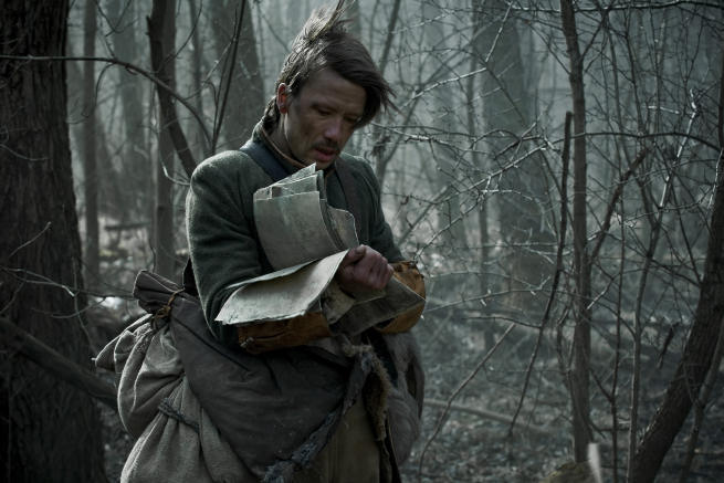 Vanhanaikaisiin vaatteisiin pukeutunut mies, jonka tukkaa tuuli tuivertaa, seisoo keskellä aavemaista kylmää korpea sylissään läjä vanhanaikaisia paperiarkkeja.