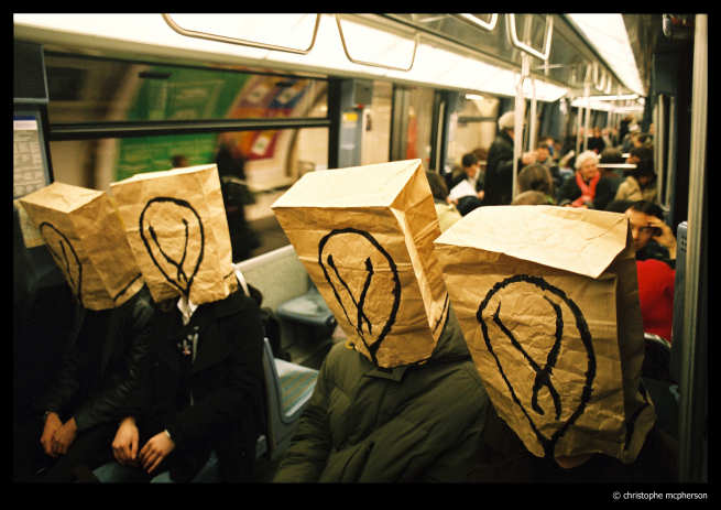 Christophe Mcphersonin valokuvassa näkyy lauma ihmishahmoja istumassa metrovaunussa. Etualalla näkyy neljän ihmisen hahmot. Heillä ruskeat pahvipussit päässä. Pussien etukylkiin maalattu mustalla tussilla hehkulamppua muistuttavat symbolit.